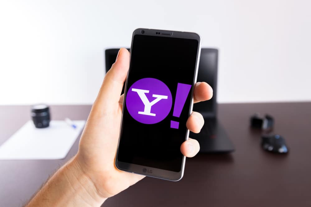 Logotipo do Yahoo é exibido na tela de um smartphone que, por sua vez, é segurado por uma mão; ao fundo, há uma mesa de escritório, com um notebook e um mouse.