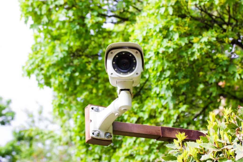 Câmera de vigilância CCTV IP branca contra a folhagem verde de uma árvore. Conceito de segurança, território privado, controle de propriedade. Programa de busca por criminosos com reconhecimento facial. Tecnologia moderna.